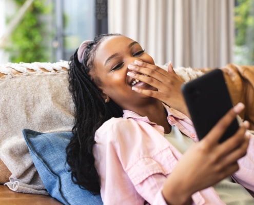 Young African American woman laughs while looking at her phone, reclining on a couch