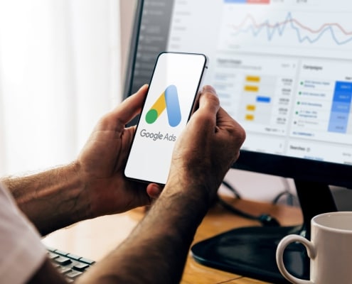 man using a mobile phone with google ads on the screen and in the background a computer showing ad statistics